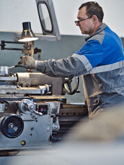 Turner in overalls works at lathe in turning shop at factory. Elderly worker turns metal part on machine. Side view.