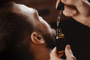 Barber applies beard oil with dropper for man in barbarshop.