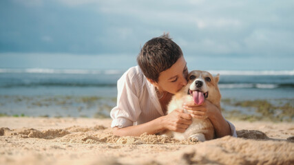 Portrait of a woman with dark hair and a short haircut kissing her Welsh Corgi dog while walking on the beach. Love and affection for your pets. Woman and corgi on the beach.