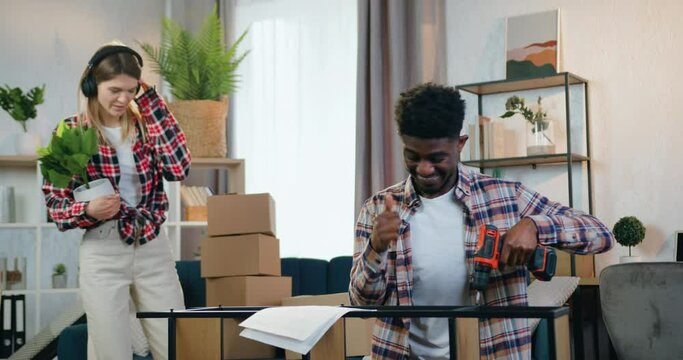 Attractive couple man and woman spending their time at home guy screwing rack with handheld screwdriver while girl enjoying music in earphones with dance