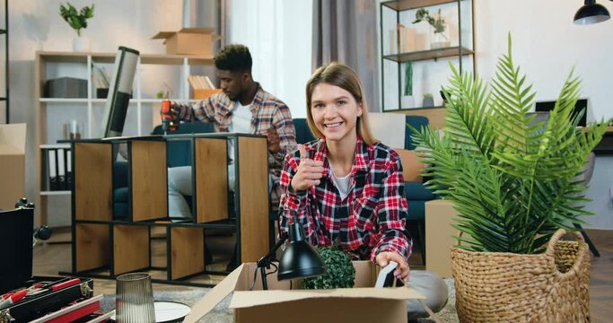 Attractive woman looking into camera with thumb up on the background of likable skillful man which assembling rack with screwdriver