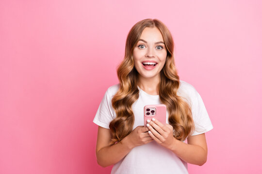 Photo of impressed cute woman with wavy hair dressed white t-shirt hold smartphone astonished staring isolated on pink color background