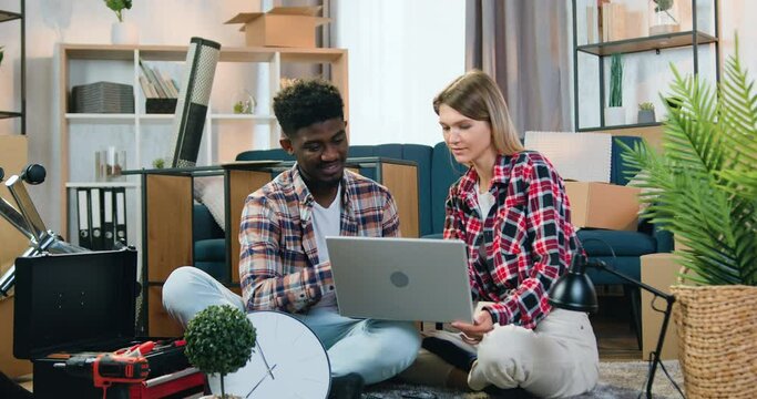 Multiethnic couple woman and man sitting on the floor among cartons tools and another things in their new apartment and use computer