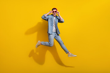Full length photo of positive excited guy wear denim jacket dark glasses jumping high isolated yellow color background