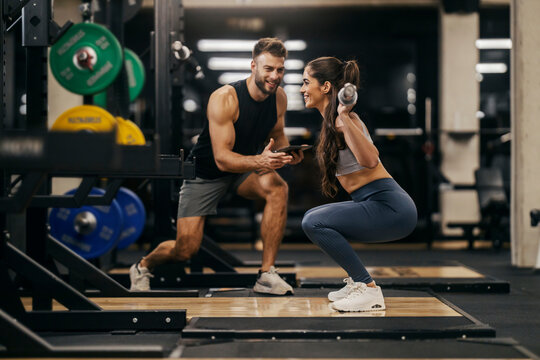 Side view of a sportswoman lifting bar in a gym with personal trainer support.