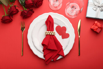 Place setting with paper hearts, gift boxes and bouquet of roses on red table, flat lay. Romantic dinner