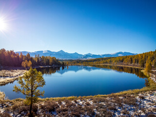 A stunning aerial shot captures a serene mountain lake with reflections of autumnal trees under a clear blue sky.