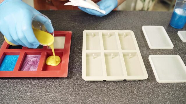 Professional craftsman in protective gloves filling silicone molds of rectangular shape with different epoxy resin colours samples, colour pigments with brilliant shadows creating exclusive natural