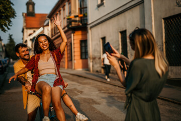 Carefree couple exploring on a bicycle, woman perched on the front wheel, another woman is taking a photo of them