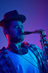 Portrait of African-American man, focused musician playing saxophone in hat against blue and pink...