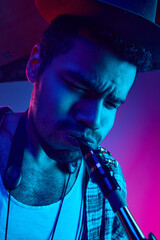 Close up portrait of young man, focused artist in retro hat playing saxophone against gradient studio background in mixed neon light. Concept of jazz, blues, classy instrumental music, concerts. Ad