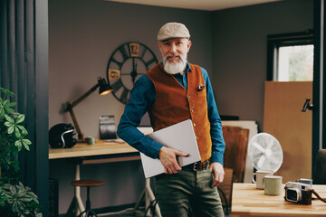 Portrait d'un homme debout souriant quinquagénaire senior hipster élégant et stylé qui fait une pause dans un atelier créatif vintage et qui porte un ordinateur portable