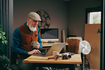Homme photographe assis souriant quinquagénaire senior hipster élégant et stylé qui travaille sur un ordinateur dans un atelier créatif vintage et qui porte un gilet et un béret  en buvant un café