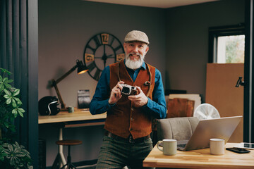 Portrait d'un photographe souriant quinquagénaire senior hipster élégant et stylé qui travaille sur un ordinateur dans un atelier créatif vintage et qui porte un gilet et un béret