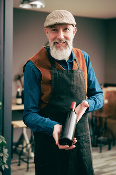 Portrait d'un sommelier vigneron vintage hispter avec un tablier et un b&eacute;ret et qui tient une bouteille de vin rouge dans une cave