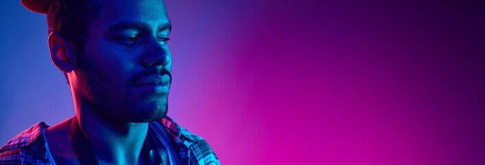 Portrait of African-American jazzman in hat looking away in neon light against gradient blue-pink...