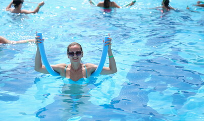 Young woman in sunglasses holding noodle for aqua aerobics in swimming pool. Sports group training on water concept