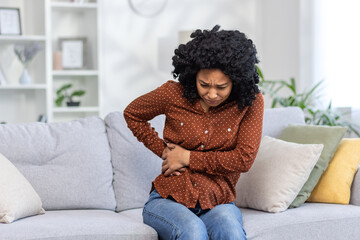 African American young woman sitting on sofa at home and holding her side of body and stomach....
