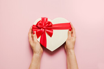 Valentine's Day romantic white heart box with red bow in woman hands on pink background. View from above. Flat lay. Greeting card with copy space.