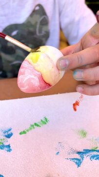 11-year-old boy watercolor painting an Easter egg red and yellow with a paint brush on a table in his backyard. Traditional kid's Easter craft. Sunny outdoor in spring.