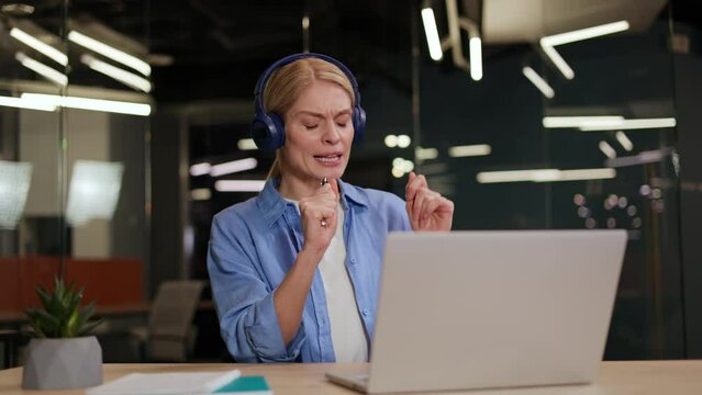 Positive young woman sitting at workdesk in front of computer and using wireless headset and pen as microphone. Active caucasian freelancer listening music and singing while having break indoors.