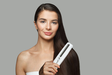 Brunette model woman holding hair iron and straightening her healthy long dark hair on white background. Haircare and hair styling concept