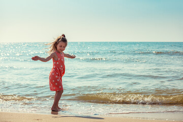 Happy little girl walking on the beach in summer. Baby girl in a pink dress with polka dots