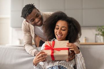 Smiling African man surprising woman with present in living room - obrazy, fototapety, plakaty