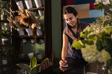 Young woman working in her flower shop talking on the phone and receiving an order for a bouquet of flowers.