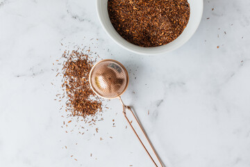 Loose leaf Rooibos tea spilling around a infuser or strainer on a marble surface with more dried tea in a white bowl