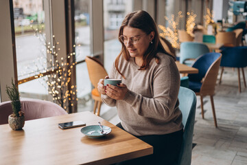Attractive female in glasses with cute smile having a coffee while relax in in a break. Positive woman in casual wear smiling and resting with cup of hot drink in cozy cafe. Concept of easy breakfast