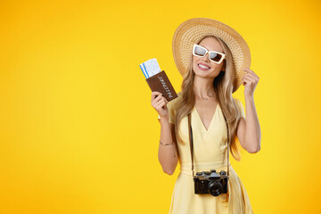 Tourist woman smiling while holding retro camera and travel tickets isolated over yellow...