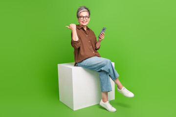 Full size photo of pleasant person hold smartphone sit on platform indicating at sale empty space isolated on green color background