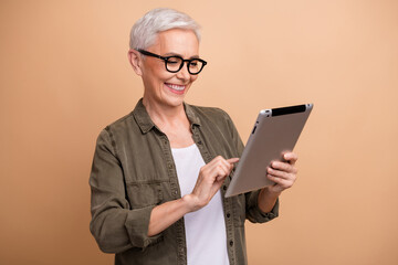 Portrait of cheerful mature curious woman wearing khaki shirt tapping tablet searching business ideas isolated on beige color background