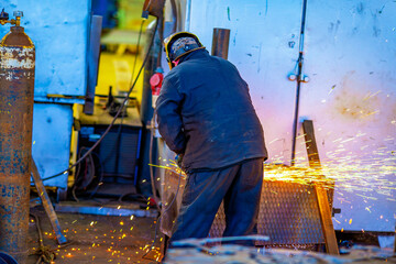 A worker cuts metal structures and parts with an abrasive tool at a factory. The grinder sparks when cutting steel. - obrazy, fototapety, plakaty