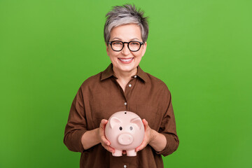 Portrait of smart optimistic senior person with short hair in eyewear hold piggy money box in arms...