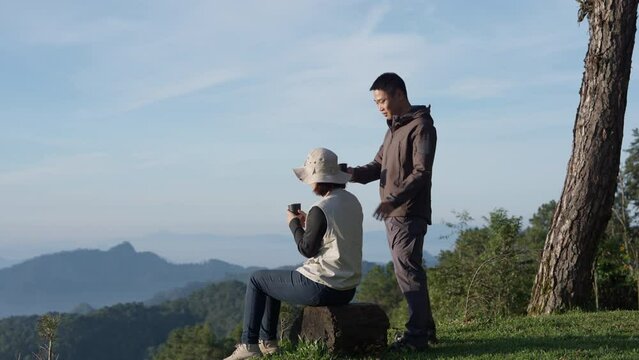 Asian happy tourist couple drinking coffee admire the mountainside scenery with hat and casual clothes in summer vacation trip on annual holiday, sun rises in the morning.