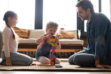 Caucasian family with their little daughters builds tower while playing on floor of children's room. Dad and two girls have fun together