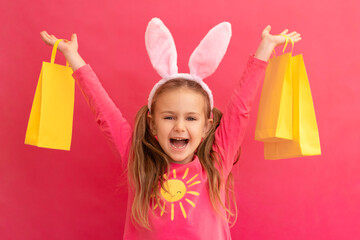 Easter shopping. Excited emotion surprise adorable child girl in bunny ears rabbit costume hold yellow shopping bags isolated pink background with finger pointed up. Kid Sale discounts easter children
