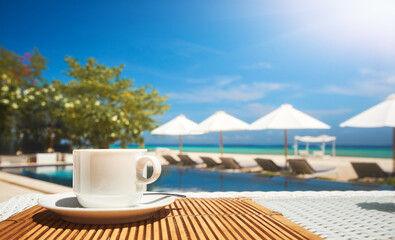 Coffee cup on table in restaurant on beach against backdrop of swimming pool and beach umbrellas on summer day. Focus on table with coffee cup.
