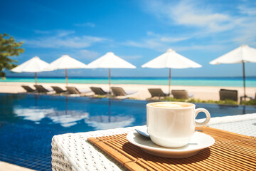 Coffee cup on a table in a restaurant on the beach against the backdrop of a swimming pool and beach umbrellas on a summer day. Focus on the table with a coffee cup. - 724738132