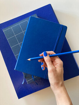 Hand with blue nail polish, holding a blue pencil on a stack of blue books, notebooks and folders.