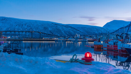 Peaceful evening at Tromsø harbor with calm waters reflecting boats and snow-clad mountains,...