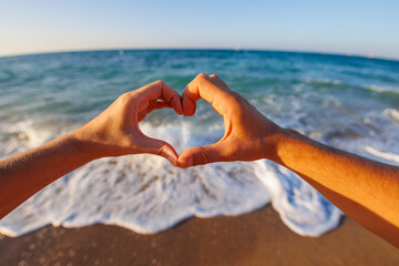 happy young couple showing heart shape with heart shaped hands on sea background, sign at sunrise over sea beach.