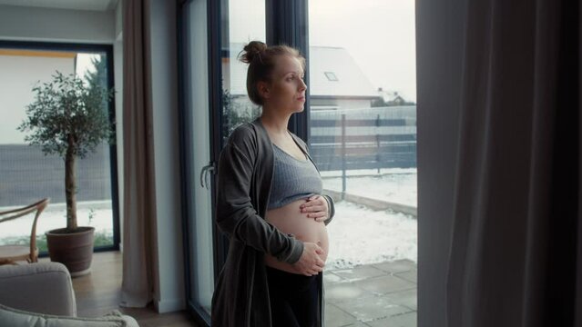 Anxious pregnant woman standing next to the window and touching her abdomen