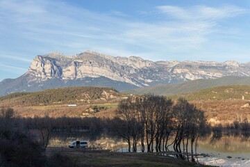 Majestic mountain range towers over a calm lake with reflections of leafless trees and a solitary van parked nearby, Aragon, Spain