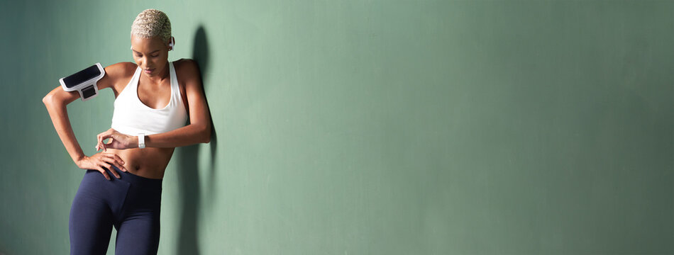 sport female athlete running training and jogging, smiling African American woman look fitness smartwatch, wears earphones and smartphone on armband. Leaning against a green urban wall with copy space