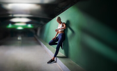 sport female athlete running training and jogging, African American woman look fitness smartwatch, wears earphones and armband. Leaning against green urban wall of hallway underground with copy space