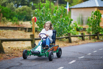Preschooler girl driving pedal race car in amusement park with road surface marking