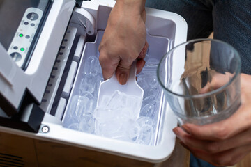 Closeup shot of a person's hand transferring bullet type ice cubes from a small portable ice maker into a clear glass with a small scoop. - obrazy, fototapety, plakaty